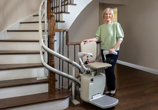 A woman standing beside her twin-rail curved stairlift on a wooden staircase