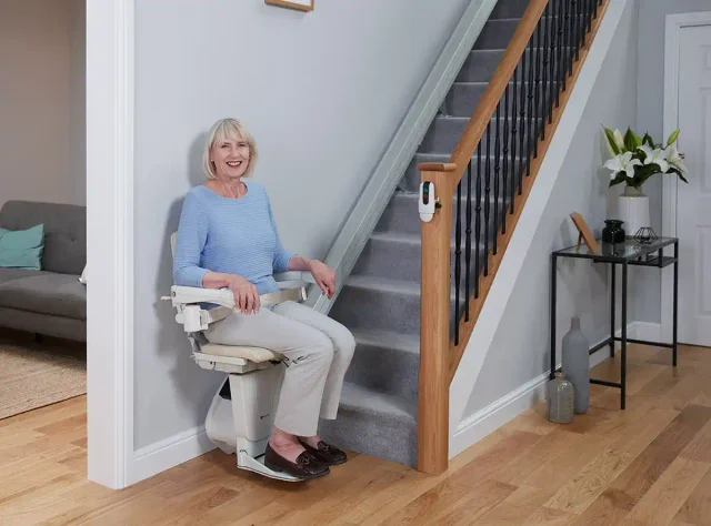 A woman seated comfortably on her stairlift at the foot of a carpeted staircase